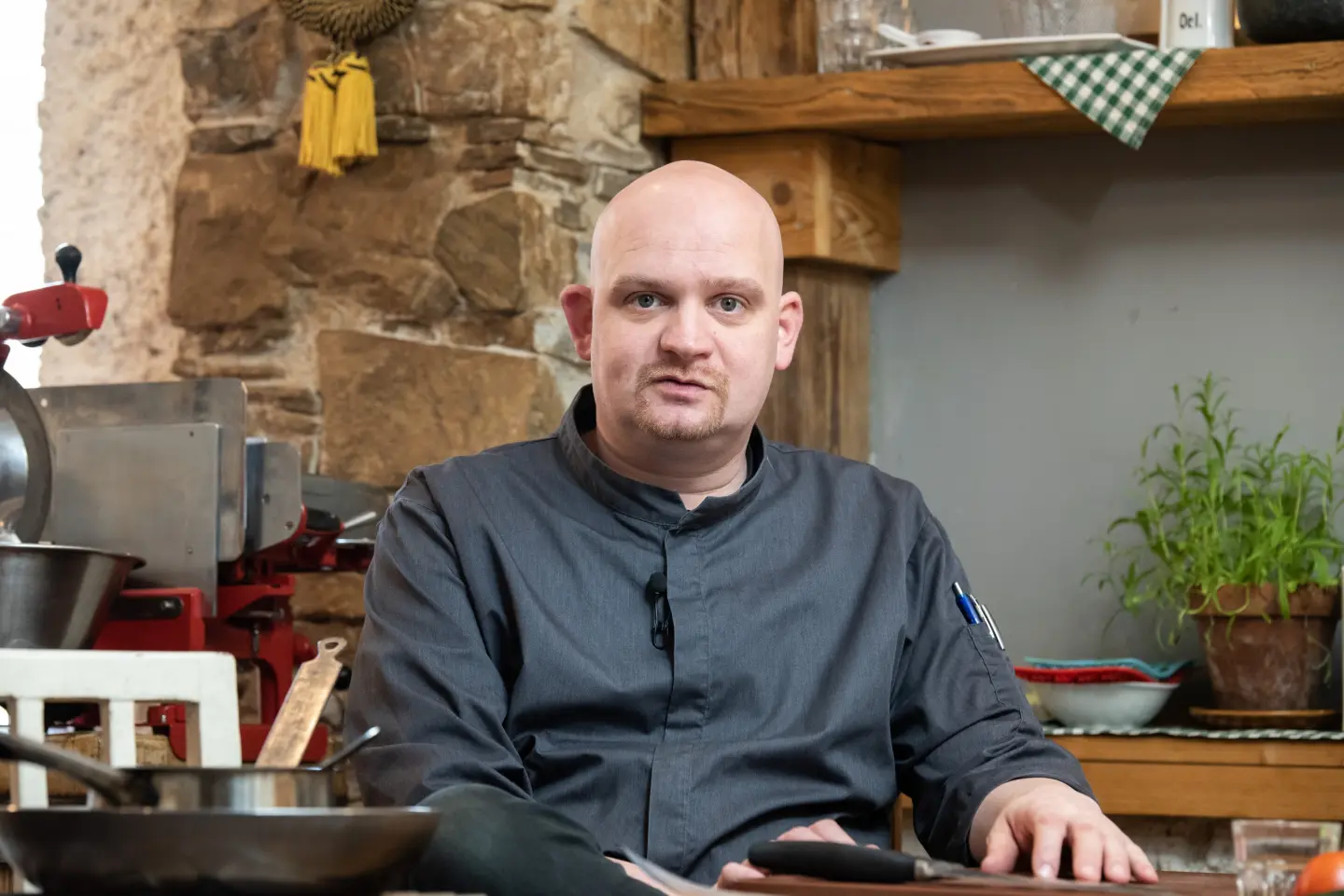 Ein glatzköpfiger Mann in einer dunklen Kochjacke sitzt in einer rustikalen Küche mit Steinwänden, Holzregalen und Kräutertöpfen und blickt direkt in die Kamera. Um ihn herum sind Kochutensilien und Küchengeräte zu sehen.