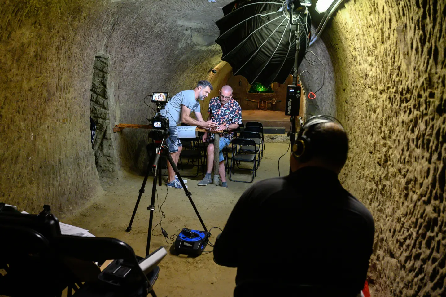 In einem unterirdischen, tunnelartigen Raum mit rauen, erdigen Wänden sitzen zwei Männer und unterhalten sich unter Studiolicht und Kameraausrüstung. Eine dritte Person mit Kopfhörern überwacht die Aufnahme im Vordergrund.