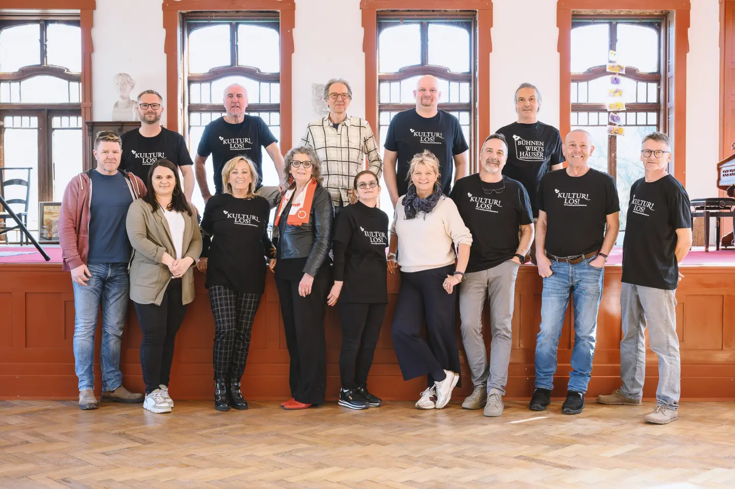 Eine Gruppe von vierzehn Erwachsenen, von denen einige passende schwarze KULTUR im LOCK-T-Shirts tragen, stellt sich für ein Gruppenfoto in einem Innenraum vor großen Fenstern und einer roten Bühne auf.