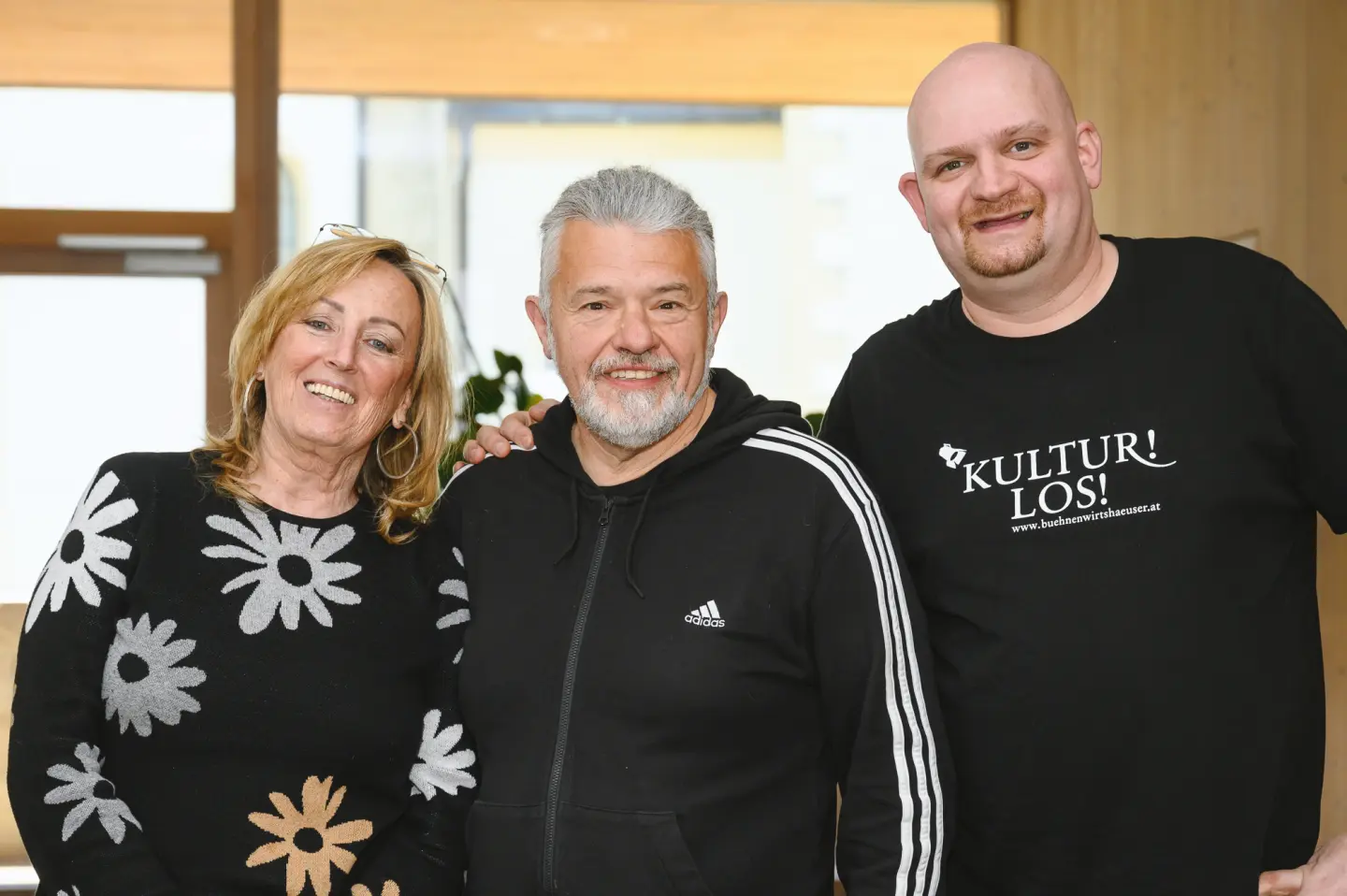 Drei Erwachsene stehen in einem Haus und lächeln in die Kamera. Die Frau auf der linken Seite trägt einen schwarzen Pullover mit weißen Blumen, der Mann in der Mitte einen schwarzen Adidas-Kapuzenpulli und der Mann auf der rechten Seite trägt ein schwarzes KULTUR! LOS!-T-Shirt.