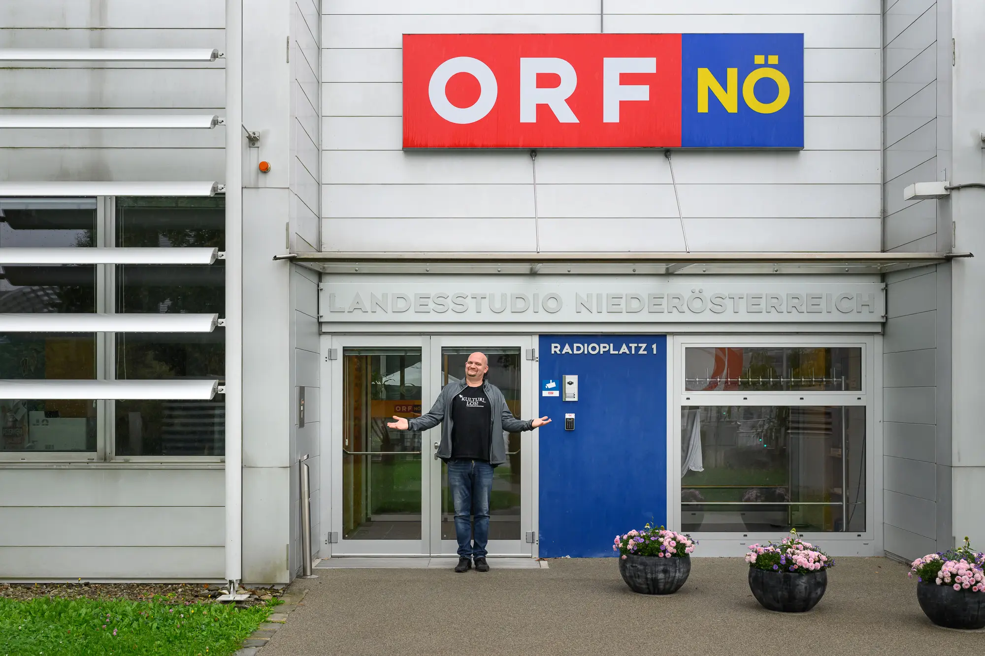 Eine Person steht lächelnd mit ausgebreiteten Armen vor dem Eingang des ORF Niederösterreich Studiogebäudes, das mit einer blauen Tür, Glasscheiben, Blumentöpfen und dem ORF NÖ Schriftzug darüber ausgestattet ist.