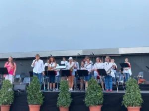 Eine Gruppe von Musikern steht auf einer Außenbühne mit Mikrofonen und Notenständern. Einige halten Instrumente in der Hand, und alle sind leger gekleidet. Topfpflanzen säumen den vorderen Teil der Bühne unter einem klaren Himmel.