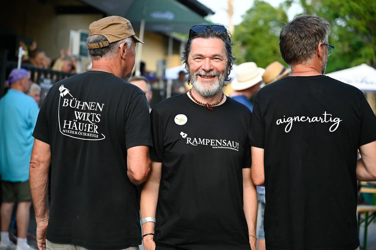 Drei Männer stehen im Freien bei einer Veranstaltung und tragen schwarze T-Shirts mit einem deutschen Text auf dem Rücken. Der Mann in der Mitte blickt lächelnd nach vorne, während die anderen beiden den Rücken zeigen. Im Hintergrund sind Menschen und Grünflächen zu sehen.