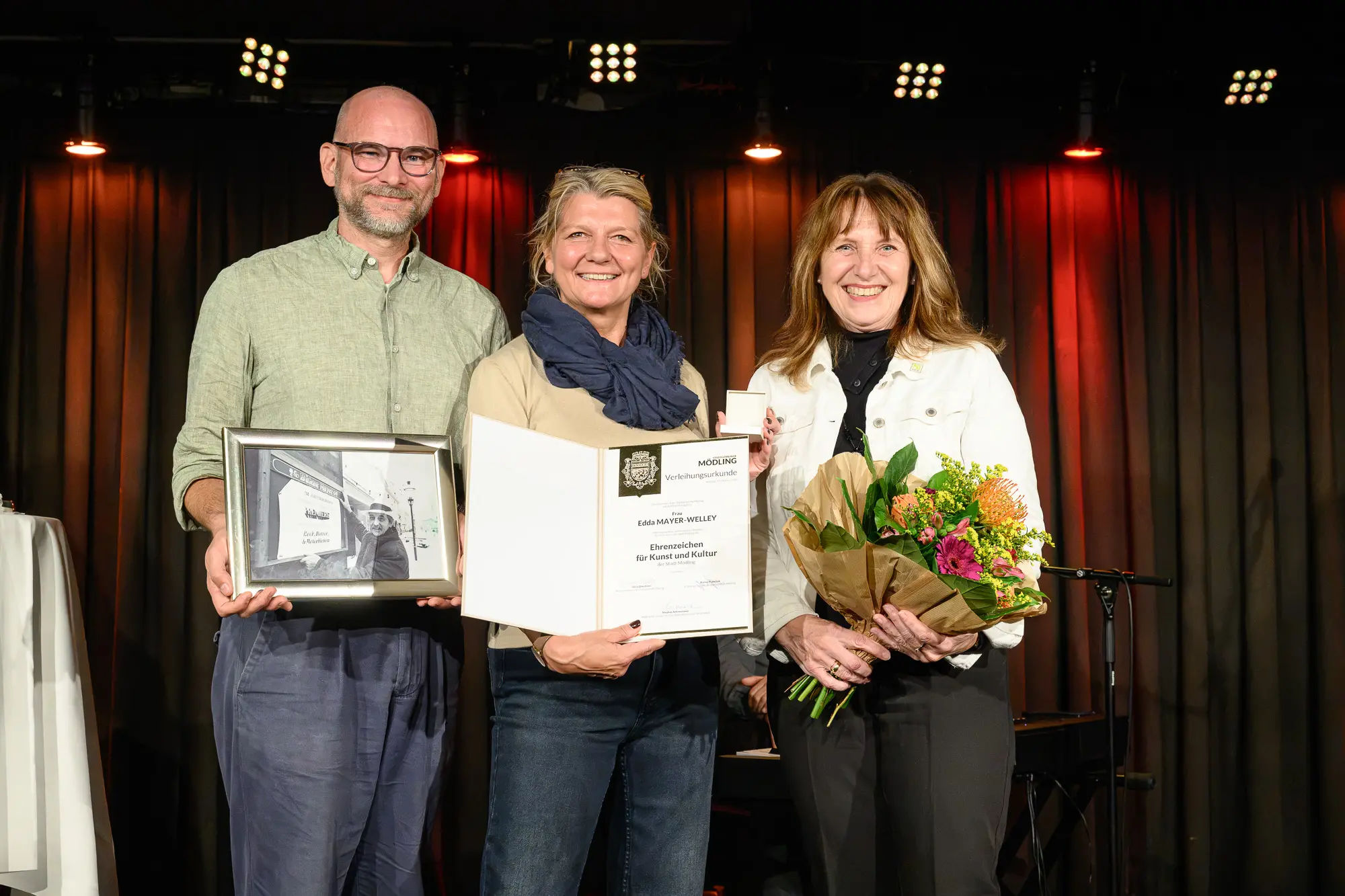 Drei Personen stehen auf einer Bühne; die Frau in der Mitte hält eine Urkunde in der Hand, der Mann links hält ein gerahmtes Foto in der Hand und die Frau rechts hält einen Blumenstrauß in der Hand.