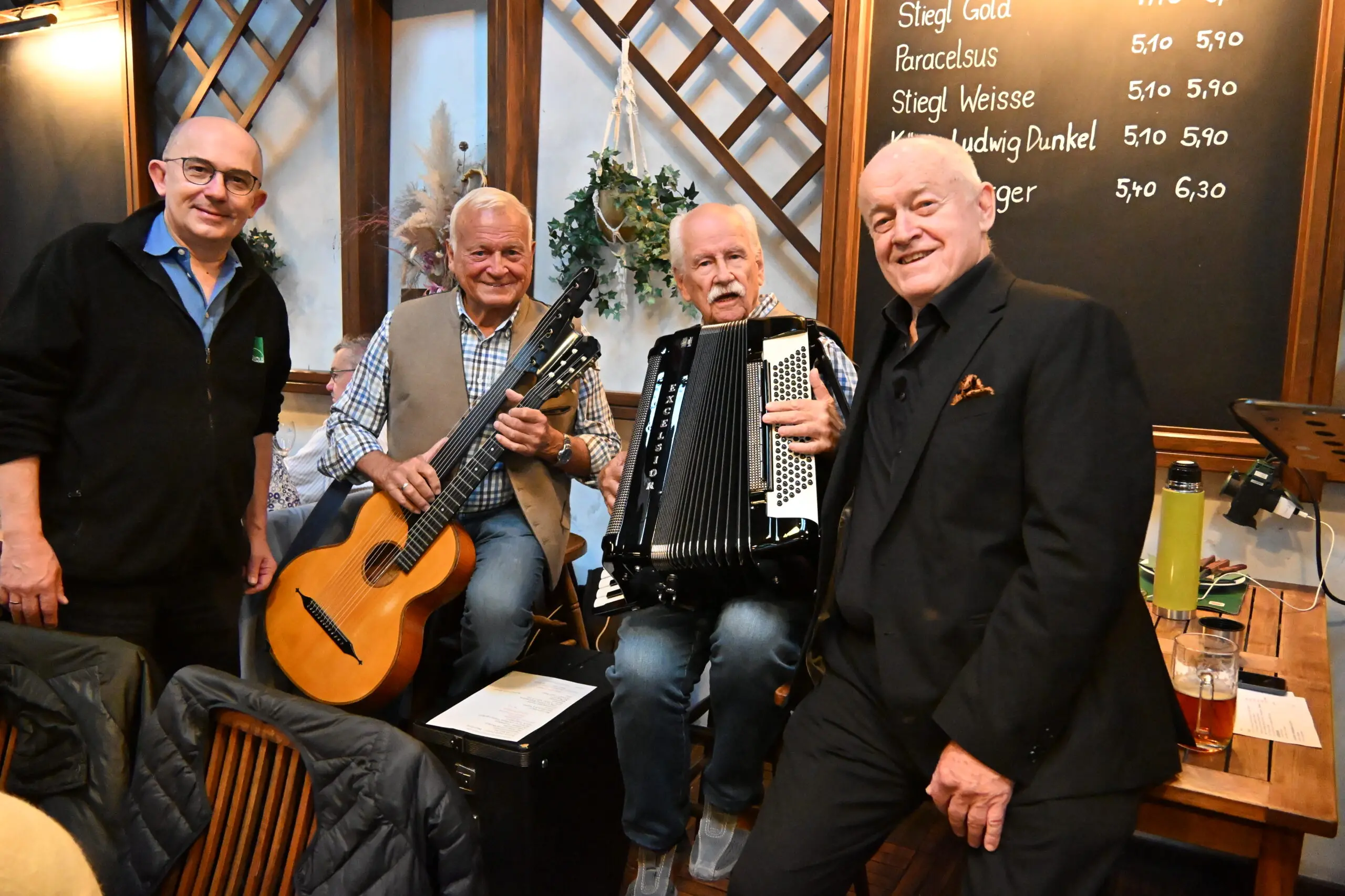 Vier ältere Männer posieren in einem Raum; zwei halten Musikinstrumente in der Hand (eine Gitarre und ein Akkordeon), während die anderen daneben lächeln. Auf dem Tisch steht ein Getränk, und im Hintergrund ist eine Kreidetafel mit einer Bierkarte zu sehen.